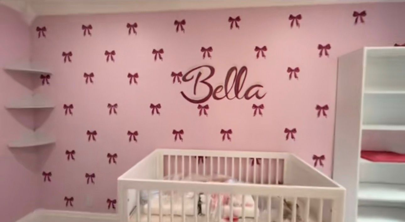 Pink nursery mural with hand-painted burgundy bows and the name Bella in script above a white crib.